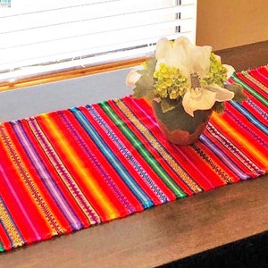 May include: A colorful striped table runner with red, orange, yellow, green, blue, and purple stripes. The runner is on a wooden table with two black chairs.