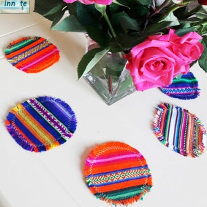 May include: Five colorful woven coasters with fringed edges. The coasters feature a variety of geometric patterns in shades of orange, blue, green, pink, and white.