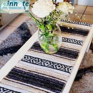May include: A white table runner with black and white stripes and a geometric pattern. The runner is made of woven fabric and is displayed on a white table with a vase of white flowers.