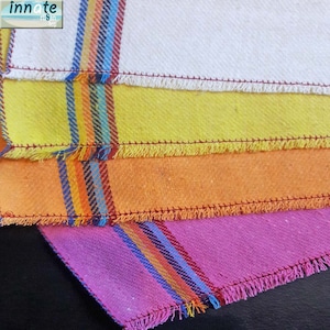May include: Three kitchen towels with fringed edges. The top towel is white with a yellow stripe, the middle towel is orange with a yellow stripe, and the bottom towel is pink with a yellow stripe. All three towels have a multi-colored stripe.