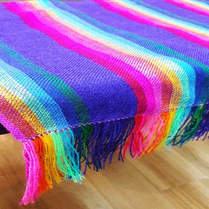 May include: A vibrant, handwoven textile with a striped pattern in shades of purple, pink, orange, yellow, green, and blue. The fabric has a fringed edge, adding texture and visual interest. The textile is likely a blanket or throw.
