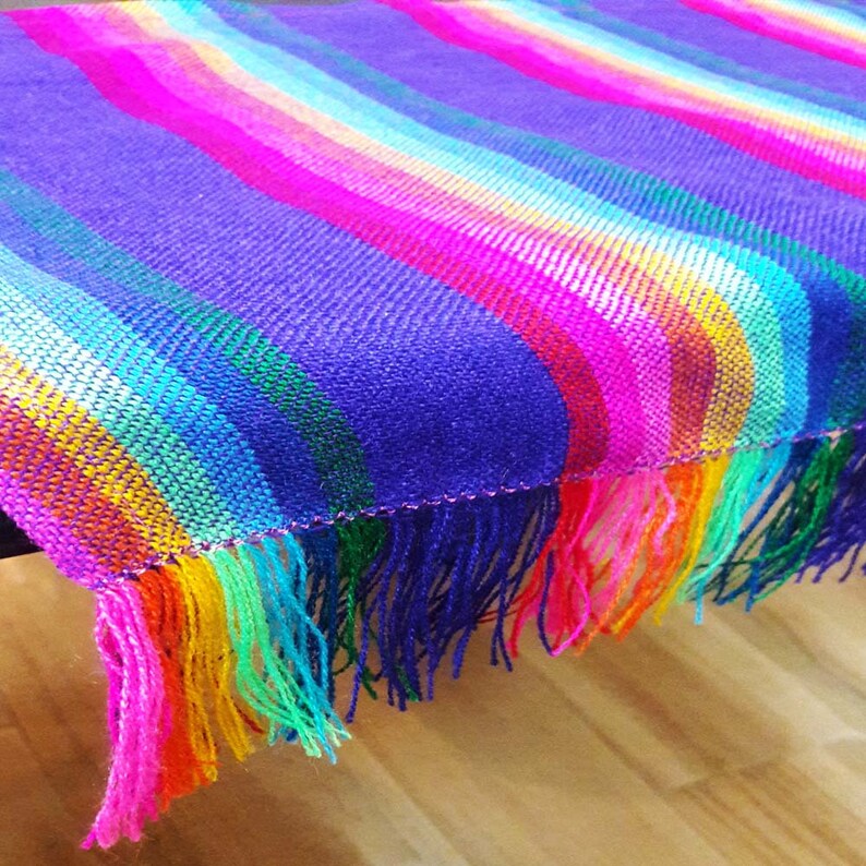 May include: A vibrant, handwoven textile with a striped pattern in shades of purple, pink, orange, yellow, green, and blue. The fabric has a fringed edge, adding texture and visual interest. The textile is likely a blanket or throw.