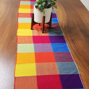 Puede incluir: Un camino de mesa a cuadros de arcoíris colorido con una planta verde en una maceta blanca sobre una mesa de madera.