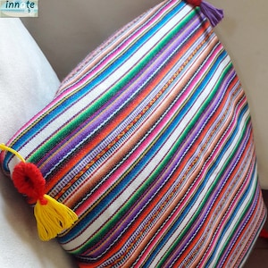 May include: Multicolored striped pillow with tassels. The pillow is made of woven fabric with a white background and colorful stripes. The tassels are yellow and red.