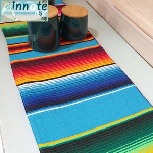 Puede incluir: Un corredor de mesa mexicano colorido con rayas verdes, amarillas, azules, blancas, negras, rojas y naranjas. El corredor está hecho de tejido tejido y tiene un borde con flecos.