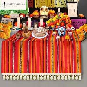 May include: A vibrant red, orange, and yellow striped table runner with a white tassel fringe. The runner is part of a Day of the Dead altar, with sugar skulls, flowers, candles, and traditional bread.