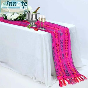 May include: A pink table runner with a colorful floral pattern and fringe. The runner is on a white tablecloth with a silver ice bucket, two wine glasses, and a single red rose.