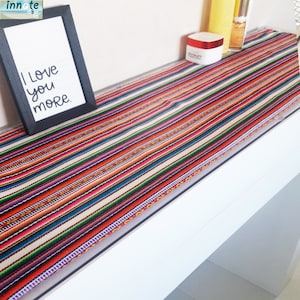 Puede incluir: Un camino de mesa a rayas de colores con rayas rojas, naranjas, amarillas, verdes, azules, moradas y blancas. El camino de mesa está en una estantería blanca con un marco de imagen que dice "Te amo más".