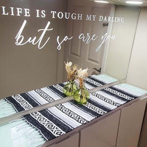 May include: A decorative mirror with the text "LIFE IS TOUGH MY DARLING but so are you". Below the mirror is a black, white, and gray patterned runner with fringe. Two small green glass vases hold dried flowers.