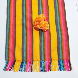 May include: A colorful, striped table runner with a yellow base and vertical stripes in pink, red, teal, and blue. The runner has a fringed edge and is adorned with two orange pom-poms.