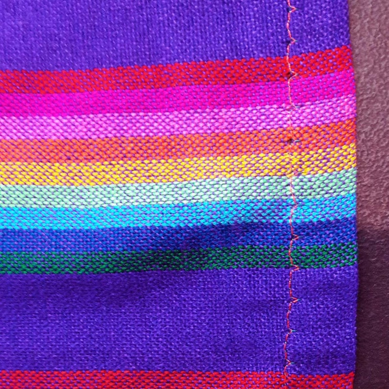 May include: Close-up of a vibrant, woven textile with horizontal stripes in a rainbow of colors, including purple, red, pink, orange, yellow, green, and blue. The fabric has a textured appearance and a visible seam with red stitching.