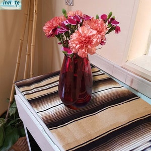 May include: A red glass vase filled with pink and peach carnations. The vase sits on a white table covered with a striped brown, black, and tan textile. Bamboo stalks are visible in the background.