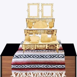 May include: A three-tiered wooden display stand featuring intricate laser-cut designs of skulls, flowers, and the text Dia de los Muertos. The stand includes two ornate picture frames at the top, a teacup, and candles, all resting on a striped table runner.