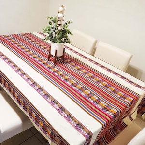 May include: A white tablecloth with colorful stripes and geometric patterns. The tablecloth is laid on a table with two chairs. A small potted plant with white cotton flowers is on the table.