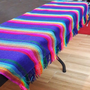 May include: A vibrant, striped textile in shades of purple, pink, orange, yellow, and green, with fringed edges. The colorful fabric is draped over a dark surface, showcasing its woven texture and bright hues. The textile is a decorative table runner.