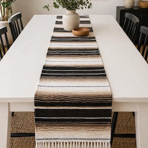May include: A woven table runner with a striped pattern in shades of black, brown, and cream, with a fringed edge. The runner is displayed on a white table, with a vase and wooden bowl on top. The table is set in a dining room.