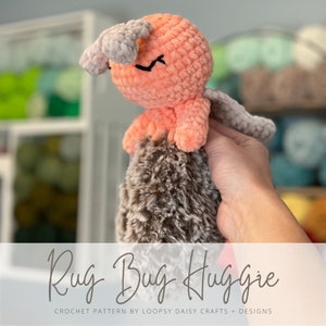 May include: A crocheted rug bug huggie toy in pink and brown. The toy has a fuzzy brown body and a pink head with a smiling face. The toy is being held by a hand. The text "Rug Bug Huggie" is written below the toy.