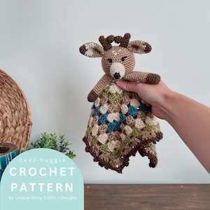 May include: A crocheted deer huggie with a brown body, antlers, and ears. The deer's face is beige with a black nose. The blanket portion is a patchwork of green, blue, beige, and brown. The text "deer huggie CROCHET PATTERN" is visible.
