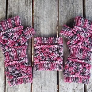 May include: A pair of pink and gray fingerless gloves with a textured knit pattern. The gloves have a small pom-pom on the top of each finger.