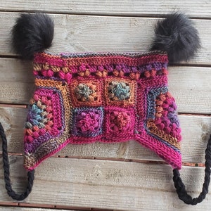Puede incluir: Un gorro de ganchillo hecho a mano con un diseño de patrón cuadrado. El gorro presenta una vibrante mezcla de hilos rosas, morados, naranjas y azules. Tiene dos pompones negros en la parte superior y lazos trenzados negros.