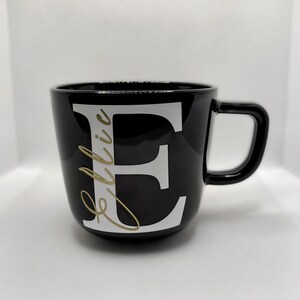 Può includere: Tazza in ceramica nera con una lettera "E" bianca e il nome "Ellie" scritto in caratteri dorati.