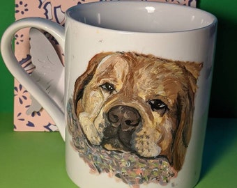 May include: White ceramic mug featuring a painted portrait of a brown dog with a colorful scarf. The handle is shaped like a white wing. A pink and blue patterned card is behind the mug.