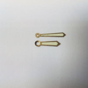 Puede incluir: Dos agujas de reloj con acabado met&aacute;lico dorado y centro rectangular blanco. Las agujas tienen un extremo puntiagudo y un agujero circular en el extremo opuesto. Las agujas son probablemente para un reloj.