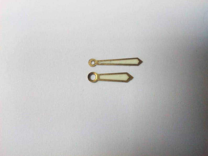 Puede incluir: Dos agujas de reloj con acabado met&aacute;lico dorado y centro rectangular blanco. Las agujas tienen un extremo puntiagudo y un agujero circular en el extremo opuesto. Las agujas son probablemente para un reloj.