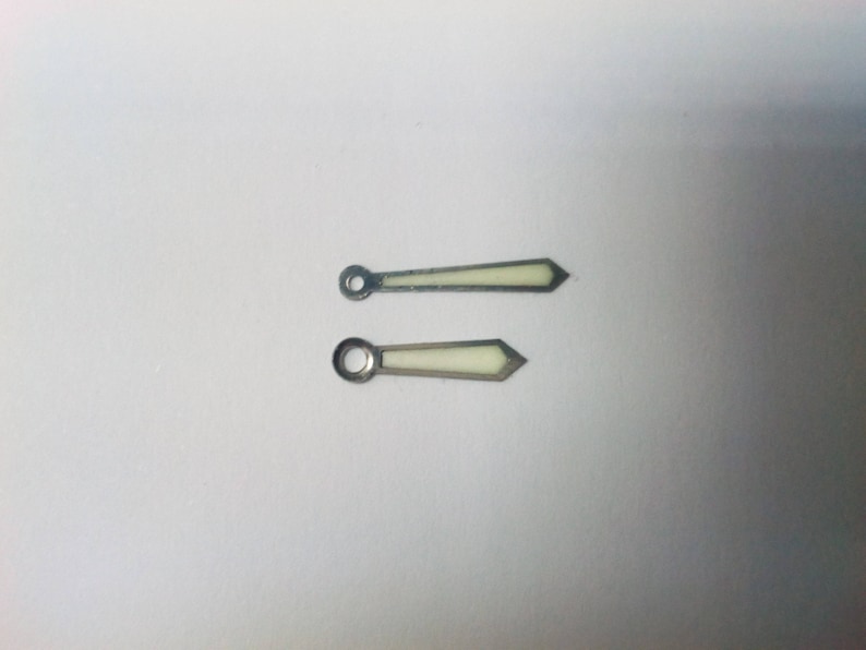 Puede incluir: Dos agujas de reloj con acabado met&aacute;lico plateado. Las agujas tienen un extremo puntiagudo y una abertura circular en el extremo opuesto. El centro de las agujas es de color crema.