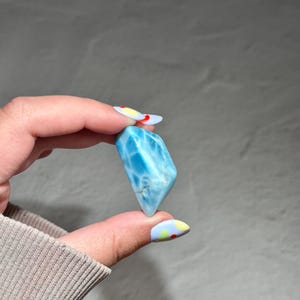 May include: A polished, light blue gemstone with white veining. The stone is held between fingers, showcasing its smooth, rounded shape and natural color variations. The gemstone is a beautiful example of natural crystal formations.