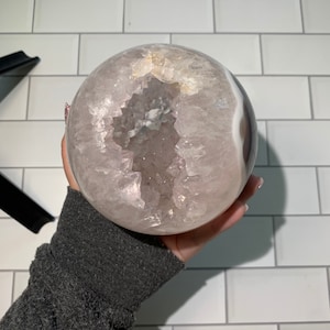 Könnte beinhalten: Eine große, kugelförmige Geode mit helllila und weißem Äußeren. Die Geode hat eine hohle Mitte, die mit glitzernden Kristallen gefüllt ist. Die äußere Oberfläche hat ein glattes, poliertes Aussehen. Die Geode wird in einer Hand gehalten.