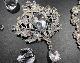 May include: A silver and crystal brooch with a large teardrop-shaped crystal in the center. The brooch is surrounded by smaller crystals and has a dangling teardrop-shaped crystal.