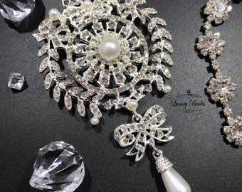 May include: A silver and white bridal brooch with a large pearl drop. The brooch features a circular design with a large pearl in the center, surrounded by smaller pearls and rhinestones. The brooch is attached to a silver ribbon bow with a smaller pearl drop.