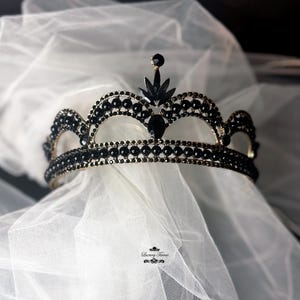 Può includere: Una corona nera e dorata con accenti di perline nere e una punta appuntita. La corona è su uno sfondo di tulle bianco.