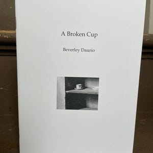 May include: A white book with the title "A Broken Cup" by Beverley Daurio. The cover features a black and white photograph of a cup on a shelf.
