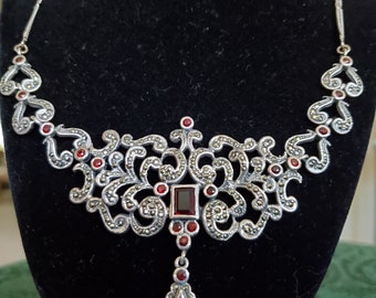 May include: A silver necklace with a filigree design and red gemstones. The necklace has a pendant with a teardrop-shaped gemstone.