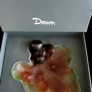 May include: A Daum France glass sculpture of a leaf and berries in a gray box. The leaf is a mix of green, orange, and red. The berries are dark gray. The box has the Daum France logo in white.