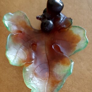 May include: A decorative leaf-shaped object with a cluster of dark, glossy berries. The leaf has a translucent appearance with shades of green, brown, and orange. The object is likely made of glass or a similar material.