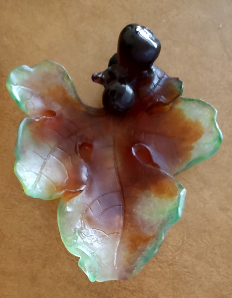 May include: A decorative leaf-shaped object with a cluster of dark, glossy berries. The leaf has a translucent appearance with shades of green, brown, and orange. The object is likely made of glass or a similar material.