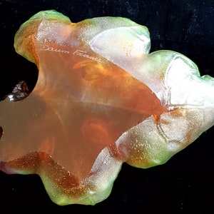May include: A translucent, leaf-shaped art glass piece with a blend of orange, green, and clear hues. The glass has a textured surface and the inscription "Daum France" is visible. The piece is set against a black background.