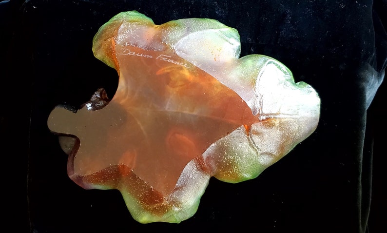May include: A translucent, leaf-shaped art glass piece with a blend of orange, green, and clear hues. The glass has a textured surface and the inscription "Daum France" is visible. The piece is set against a black background.