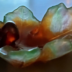 May include: A decorative glass bowl in the shape of a leaf, with a dark brown center and a translucent green and yellow rim. The bowl has a unique, organic design, perfect for displaying small items.