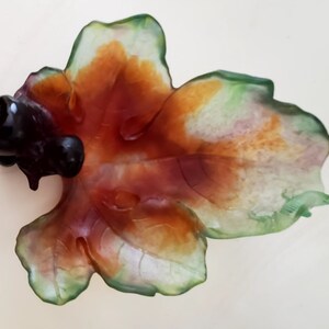 May include: A leaf-shaped decorative dish with a dark, glossy, bulbous element on the left side. The dish features a blend of colors, including green, orange, and red, with a translucent quality. The overall design suggests a natural, organic theme.