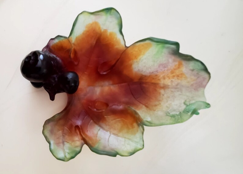May include: A leaf-shaped decorative dish with a dark, glossy, bulbous element on the left side. The dish features a blend of colors, including green, orange, and red, with a translucent quality. The overall design suggests a natural, organic theme.