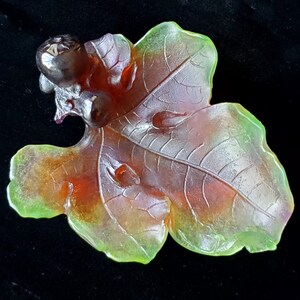 May include: A translucent glass leaf sculpture with a cluster of berries. The leaf is primarily orange and red, with green edges and detailed veining. The berries are dark purple. The sculpture is set against a black background.