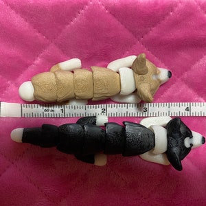 May include: Two segmented, articulated, plush dog toys. The top dog is brown and white, and the bottom dog is black and white. Both dogs are lying on a pink quilted surface with a ruler measuring 5 inches.