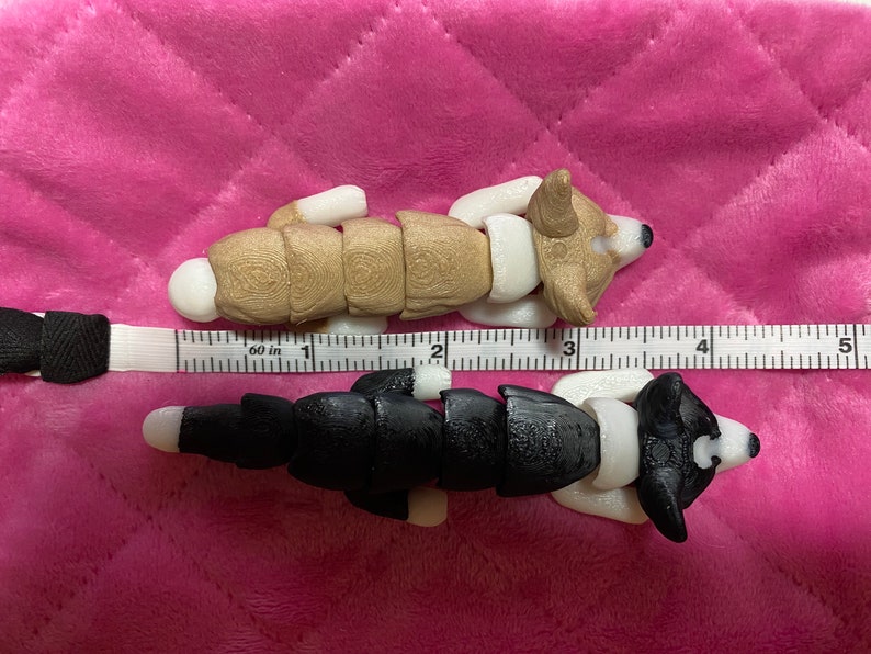 May include: Two segmented, articulated, plush dog toys. The top dog is brown and white, and the bottom dog is black and white. Both dogs are lying on a pink quilted surface with a ruler measuring 5 inches.