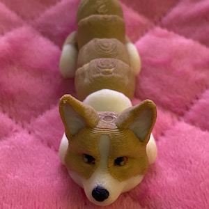 May include: A 3D printed figurine of a brown and white corgi dog lying down. The figurine is made of plastic and has a detailed design.