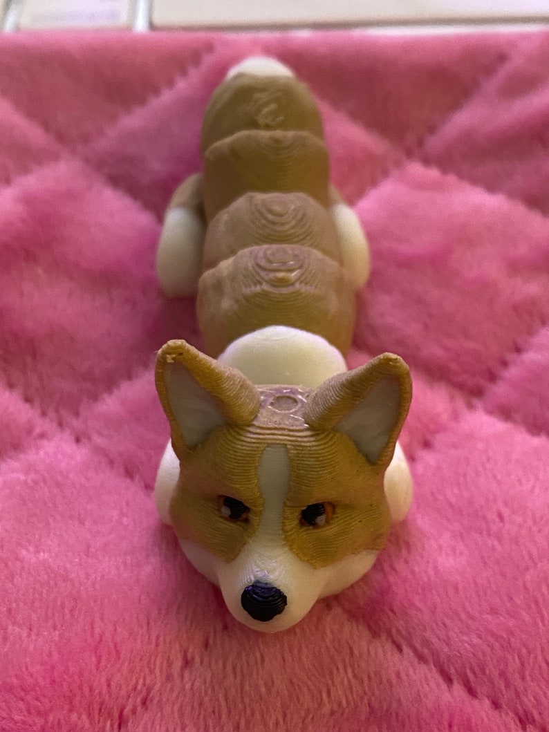 May include: A 3D printed figurine of a brown and white corgi dog lying down. The figurine is made of plastic and has a detailed design.