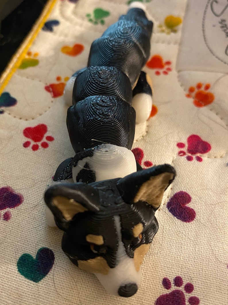 May include: A 3D printed black and white dog figurine with a curled tail. The figurine is lying on its belly with its head tilted up.
